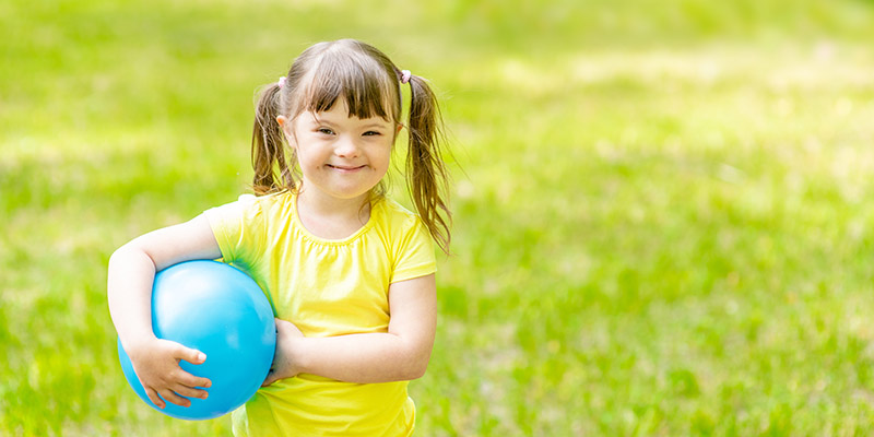 Smiling little girl with down syndrome holds ball in a park